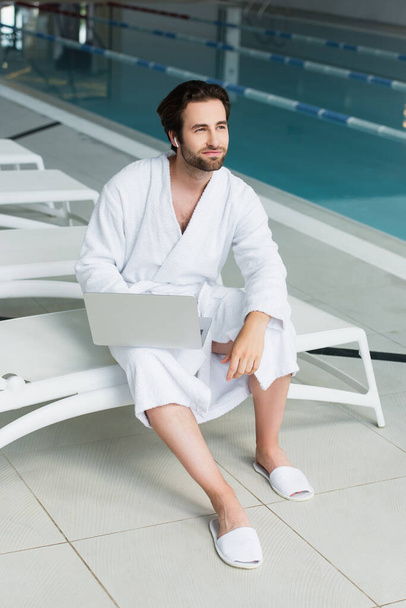 Man in bathrobe and earphone holding laptop on deck chair in spa center  - Фото, изображение