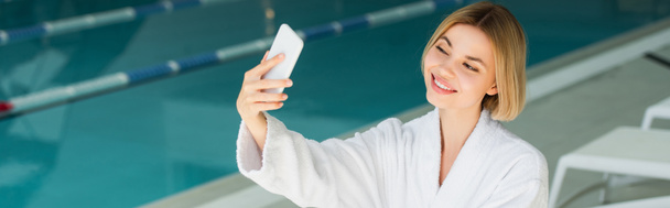 Vidám nő fürdőköpenyben csinál selfie okostelefon közelében elmosódott medence spa központ, banner  - Fotó, kép