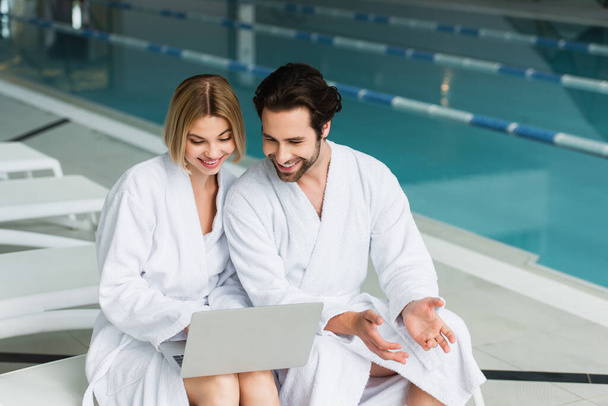 Smiling couple in bathrobes using laptop on deck chair in spa center  - Fotoğraf, Görsel