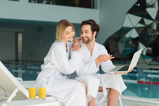 Smiling man pointing at laptop near girlfriend in bathrobe and orange juice in spa center  - Фото, изображение