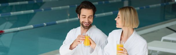 Веселая пара в белых халатах с бокалами апельсинового сока в спа-центре, баннер  - Фото, изображение