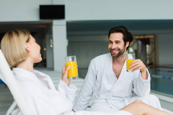 Homem sorridente em roupão segurando vidro de suco de laranja perto de namorada borrada no centro de spa  - Foto, Imagem