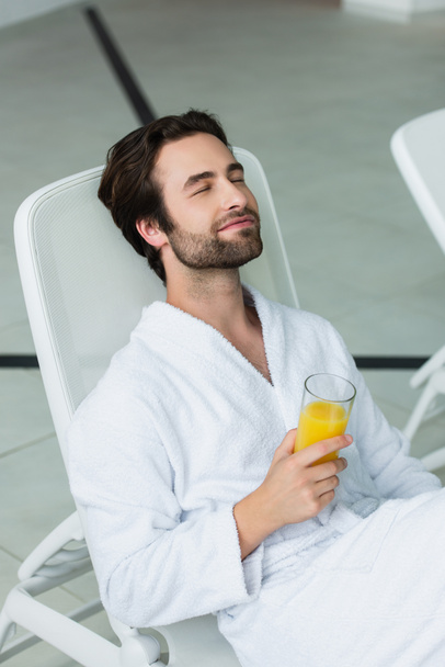 Young man in white bathrobe holding orange juice while relaxing in spa center  - Photo, Image