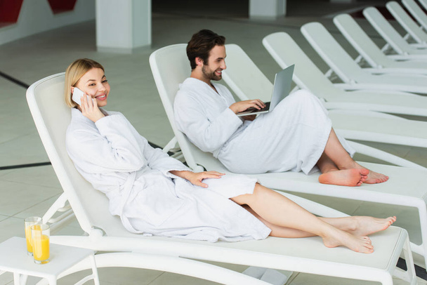 Mulher alegre falando no smartphone perto de suco de laranja e namorado com laptop no centro de spa  - Foto, Imagem