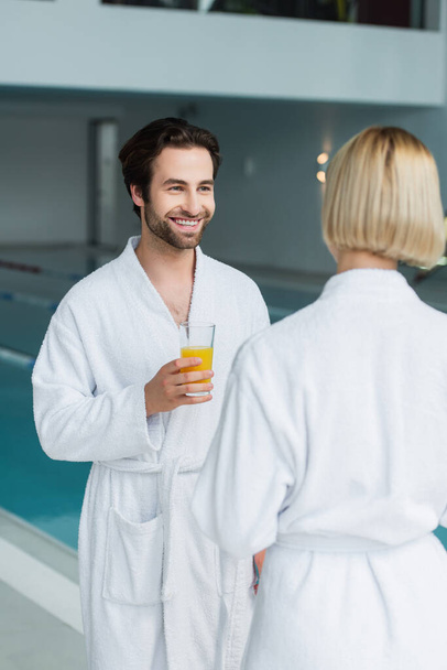 Smiling man in white bathrobe holding glass of orange juice near blurred girlfriend and swimming pool in spa center  - Фото, изображение