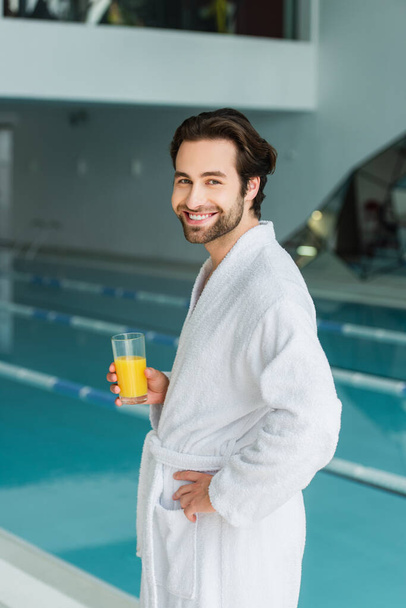 Young man in white bathrobe holding glass of orange juice near blurred swimming pool in spa center  - Foto, Bild