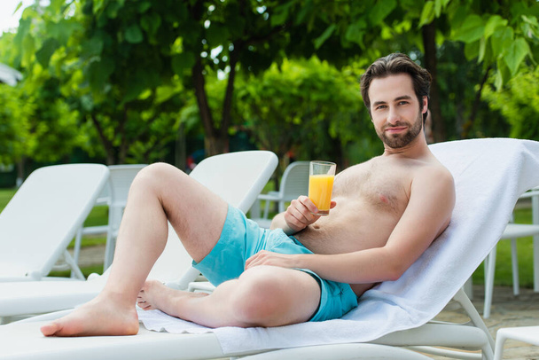 Young man in swimming trunks holding orange juice on deck chair on resort  - Photo, image