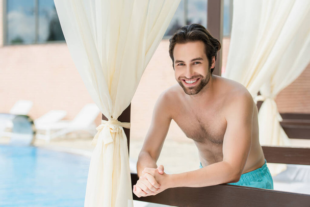 Mosolygó férfi fürdőruhában áll a lounge ágy mellett üdülőhelyen  - Fotó, kép