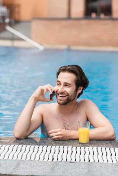 Happy man talking on smartphone near orange juice in swimming pool  - Fotó, kép
