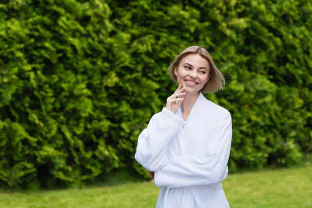 Positieve vrouw in witte badjas kijkt weg op het resort buiten  - Foto, afbeelding