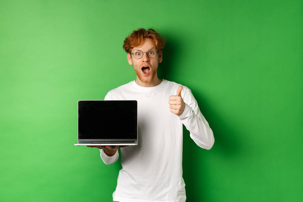 Homem caucasiano impressionado com cabelos vermelhos e barba, mostrando tela de laptop em branco e polegar para cima, olhando espantado com a câmera, elogiando algo on-line, fundo verde - Foto, Imagem