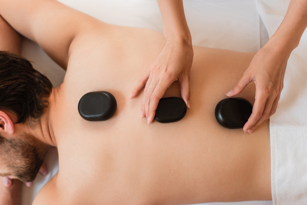 Vista ritagliata del massaggiatore mettendo pietre calde sul retro dell'uomo nel centro benessere  - Foto, immagini