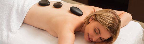Mujer joven con piedras calientes en la espalda en la mesa de masaje en el centro de spa, pancarta  - Foto, Imagen