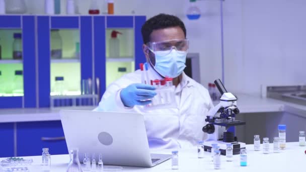 Científico indio en gafas que sostiene tubos de ensayo cerca del portátil y el microscopio  - Metraje, vídeo