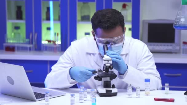 Indischer Wissenschaftler mit Brille unter dem Mikroskop in der Nähe von Impfstoffen und Laptop im Labor  - Filmmaterial, Video