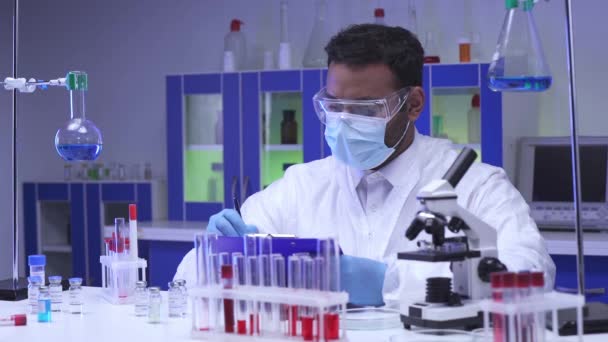 Indiase wetenschapper schrijft op klembord in de buurt van reageerbuizen en vaccins in het lab  - Video