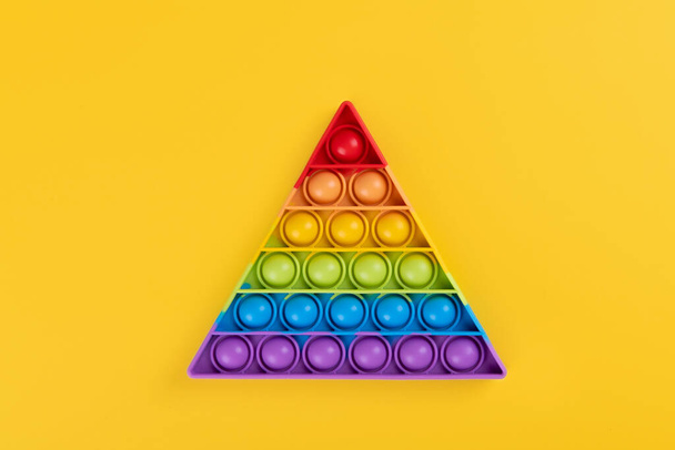 Φωτεινό ουράνιο τόξο παιχνίδι antistress για παιδιά και ενήλικες σε κίτρινο φόντο. Ευέλικτη αισθητηριακή αντι-στρες παιχνίδι ποπ το σχήμα τρίγωνο - Φωτογραφία, εικόνα