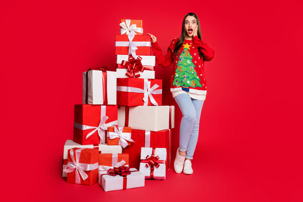 Photo of funny shocked young woman dressed ugly pullover arm cheek getting many presents isolated red color background - Foto, Bild