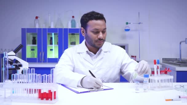 Indian scientist writing on clipboard near vaccines and test tubes in laboratory - Filmmaterial, Video