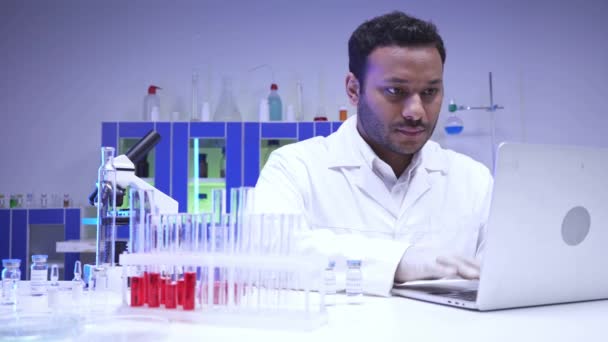 Indiase wetenschapper met behulp van laptop in de buurt van microscoop en reageerbuizen in het lab  - Video