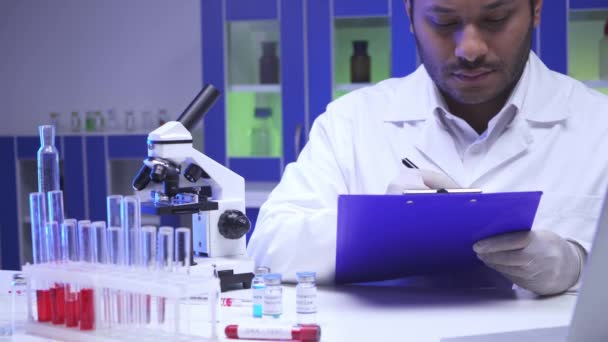 Индийский ученый пишет на планшете возле микроскопа и пробирки в лаборатории  - Кадры, видео