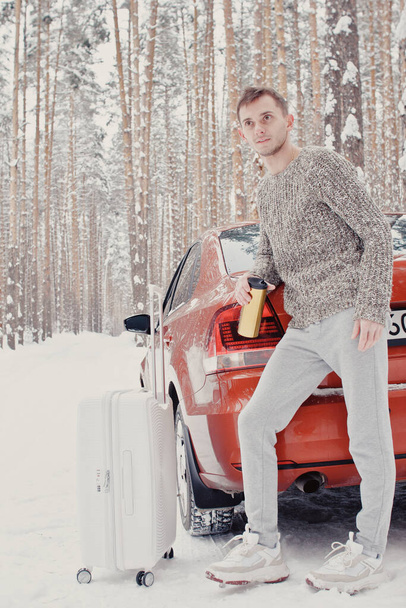 Portrait of young fashionable man in grey clothes with car on snow holding suitcase and go pathway pine forest. Winter vacation travel concept. Outdoors in winter. Ski resort, mounting skiing resort - Valokuva, kuva