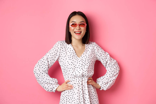 Tourismus und Lifestylekonzept. Schöne koreanische Frau im Urlaub, trägt Sonnenbrille und Kleid, sieht links mit glücklichem Lächeln, steht über rosa Hintergrund - Foto, Bild