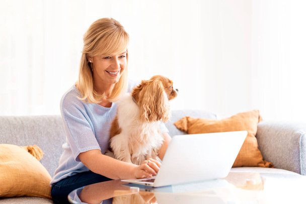 Shot of middle aged businesswoman using laptop while sitting on a couch at home and working from home. Her cute puppy relaxing in her lap. - Фото, изображение