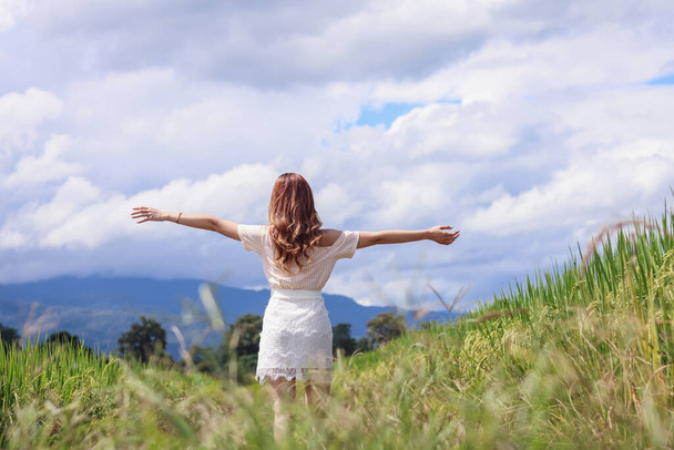 A young woman raises both her hands above her head to invoke God's blessing with the faith and power she has for God on the blurry background of a green rice field in the morning. - Photo, Image