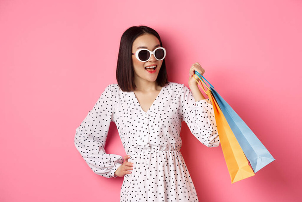 ショッピングモールに行く幸せな現代アジアの女性は、服や笑顔でバッグを保持し、サングラスを着用し、ピンクの背景の上に立って - 写真・画像