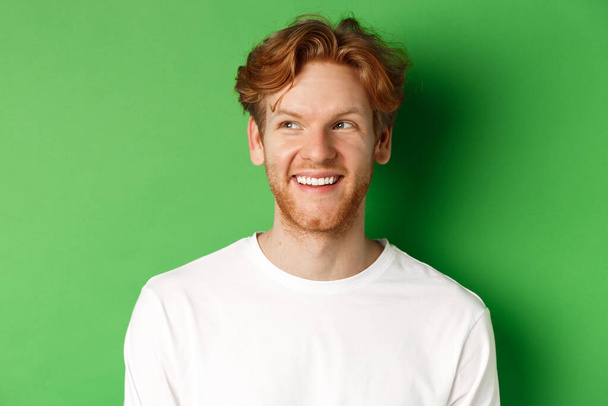 Kopfschuss eines glücklichen rothaarigen Mannes mit Bart, der weiße lange Ärmel trägt, nach links in den Kopierraum blickt und lächelt, vor grünem Hintergrund stehend - Foto, Bild