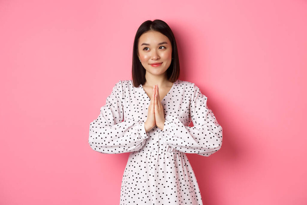 Piękna anielska Azjatka uśmiechnięta, trzymająca się za ręce w modlitwie i patrząca w lewo na przestrzeń z niewinnym uroczym spojrzeniem, stojąca nad różowym tłem - Zdjęcie, obraz