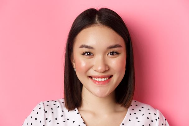 Concepto de belleza y estilo de vida. Cabeza de hermosa mujer asiática sonriendo, mirando a la cámara feliz y romántico, de pie sobre fondo rosa - Foto, Imagen