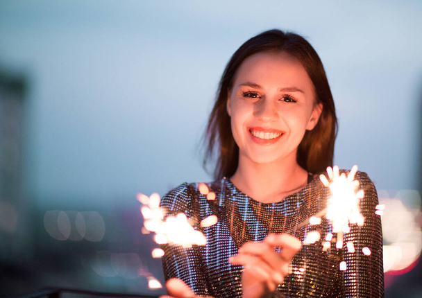 Очарованная юная леди в вечернем платье улыбается и машет горящими искрами во время празднования праздника в ночное время - Фото, изображение