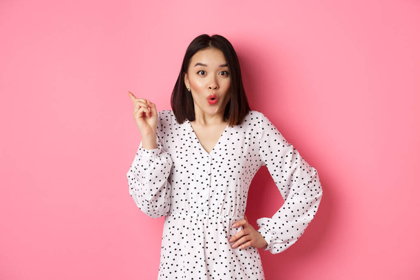 Izgalmas ázsiai női modell mutatja promo ajánlat, mutatva a bal felső sarokban, és bámulja a kamera elképedt, visel trendi tavaszi ruha, felett áll rózsaszín háttér - Fotó, kép