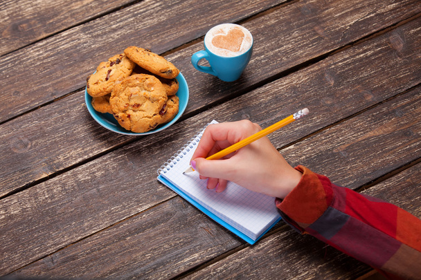 Жіноча рука пише щось в ноті біля печива і чашки кофе
 - Фото, зображення