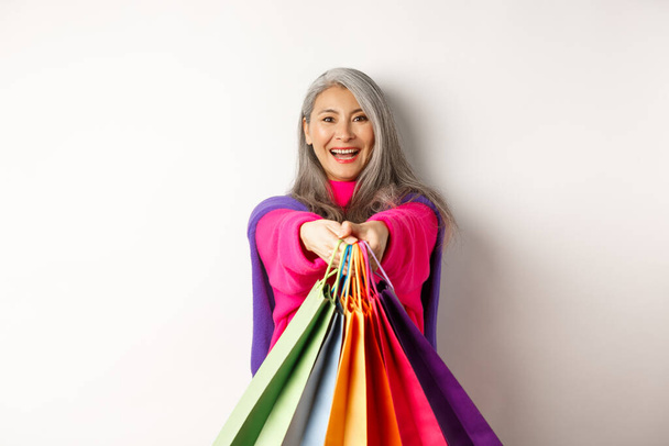 Divatos ázsiai idős nő megy vásárolni, kinyújtott kézzel papírzacskók, mosolygós elégedett a kamera, áll a fehér háttér - Fotó, kép