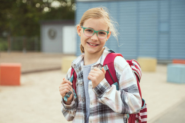 Portrait of pretty little school girl in glasses with backpack standing, posing and smiling in front of school - 写真・画像