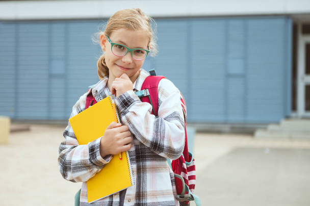 Portrait of pretty little school girl in glasses with backpack standing, posing and smiling in front of school - 写真・画像
