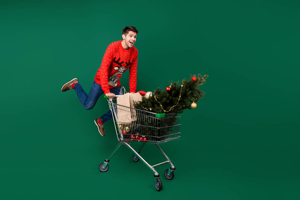 Full length body size photo man running with cart in shopping center xmas presents tree isolated green color background - Foto, immagini