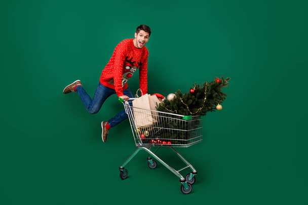 Full length photo of young excited man happy positive smile jump hold trolley shop christmas sale isolated over green color background - Fotoğraf, Görsel