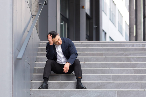 Asian businessman has a severe headache sitting on the stairs near the office - Fotoğraf, Görsel