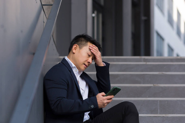 Asian businessman broker reports bad news talking on the phone, sitting near office on stairs, depressed losing hope and job bankrupt - Foto, Imagen