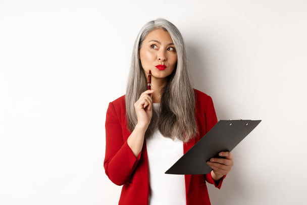 Elegante asiático oficina dama en rojo blazer escritura informe en portapapeles, buscando reflexivo mientras se trabaja, de pie sobre fondo blanco - Foto, Imagen