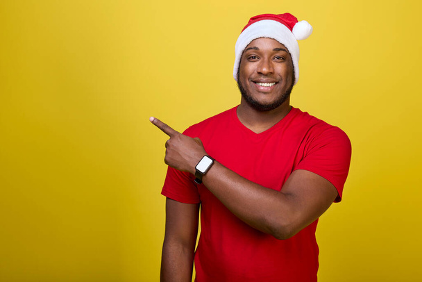 Portret van een vrolijke Afro-Amerikaanse man in een kerstman hoed met zijn wijsvinger omhoog naar een advertentie geïsoleerd op een felgele achtergrond - Foto, afbeelding