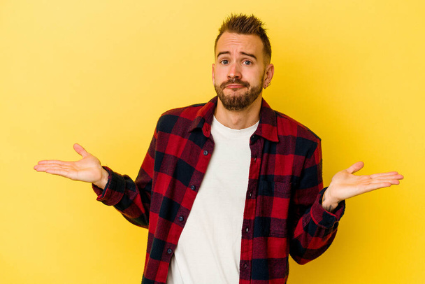 Young tattooed caucasian man isolated on yellow background doubting and shrugging shoulders in questioning gesture. - Photo, Image