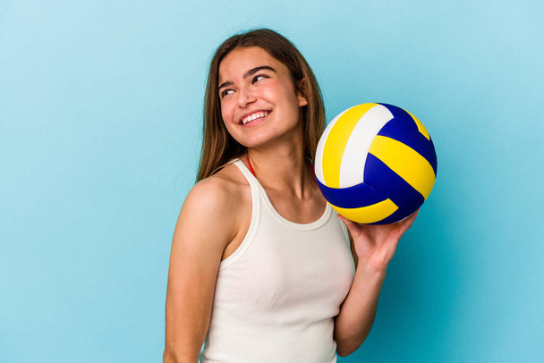 młoda kaukaska kobieta gra w siatkówkę odizolowany na niebieskim tle patrzy na bok uśmiechnięty, wesoły i przyjemny. - Zdjęcie, obraz