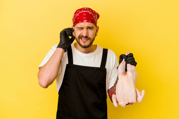 młody wytatuowany batcher kaukaski mężczyzna gospodarstwa kurczaka izolowane na żółtym tle pokrywa uszy z rąk. - Zdjęcie, obraz