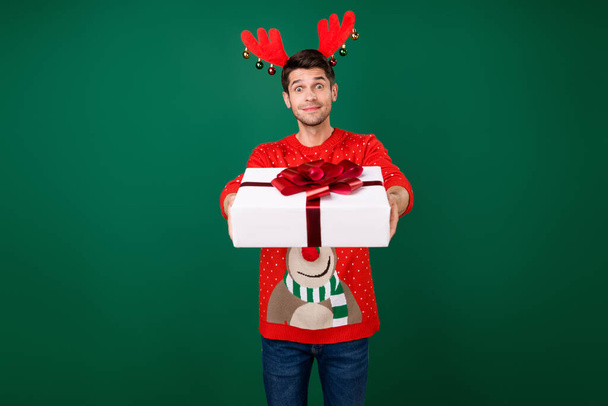 Photo of young man happy positive smile hold gift box give party wear headband reindeer isolated over green color background - Zdjęcie, obraz