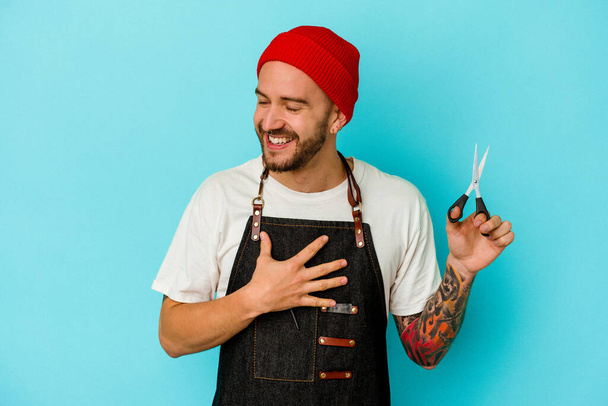 Young tattooed barber man isolated on blue background laughs out loudly keeping hand on chest. - Photo, Image
